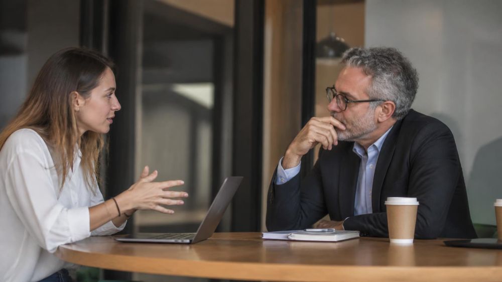 Two professionals in conversation across a table, one making a point and the other listening thoughtfully